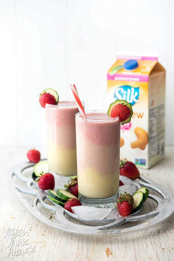 two strawberry and peach smoothies on a silver plate with cucumber strawberries and ice and a cashew milk carton in the background