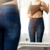 woman pulling her top up looking at her belly in the mirror