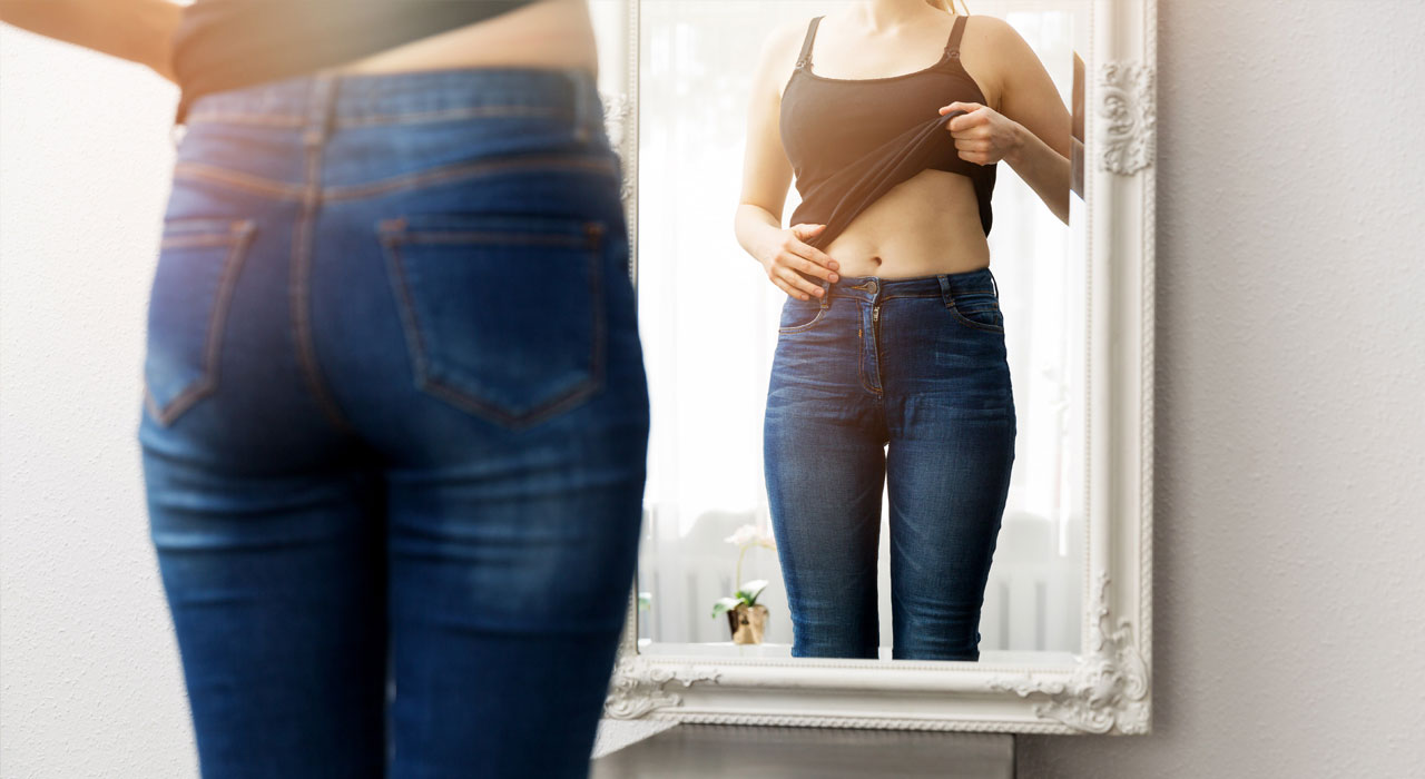 woman pulling her top up looking at her belly in the mirror