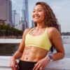 fit woman with yellow top and black gym pants leaning against railing showing abs