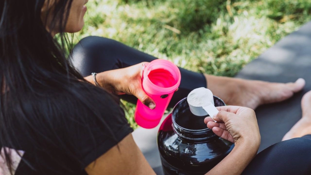 woman dressed in black gym gear scooping protein into pink protein shaker