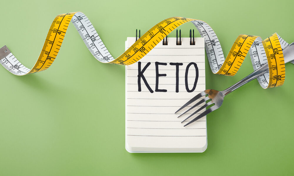 note pad with the word keto written on in capital letters and a measuring tape wrapped around a fork on a light green background