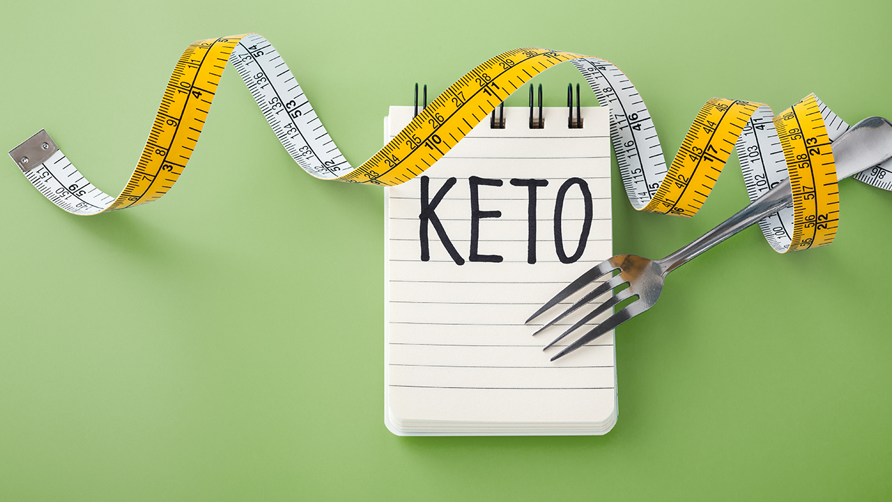 note pad with the word keto written on in capital letters and a measuring tape wrapped around a fork on a light green background